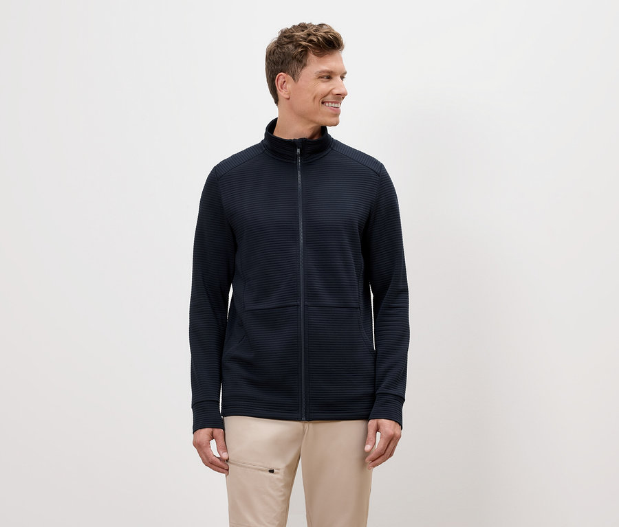 Un homme aux cheveux blonds porte une veste zippée bleu marine et un pantalon beige.