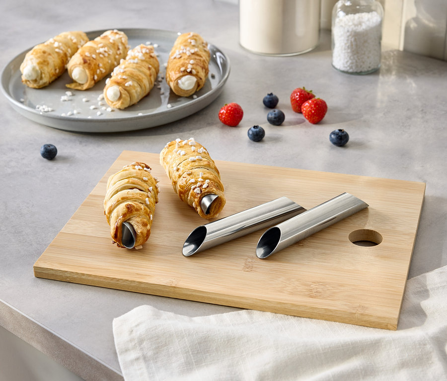 Sur une table, des cornets à la crème sont disposés sur une assiette et une planche à découper, accompagnés de baies, de sucre et de moules en métal.