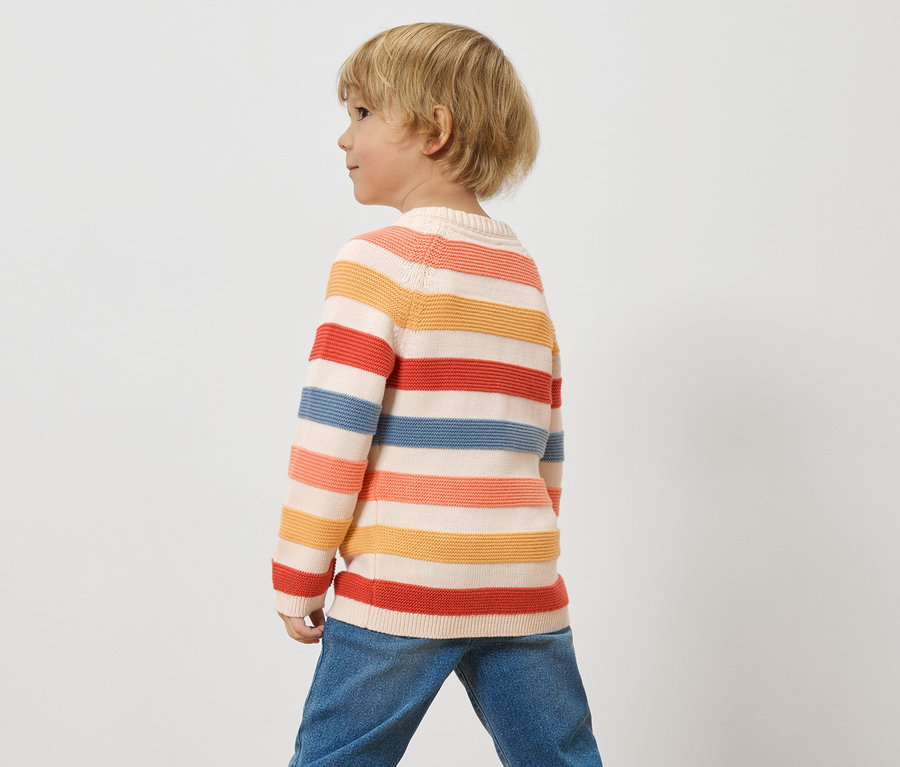Un enfant aux cheveux blonds porte un pull rayé et un jean sur un fond blanc.