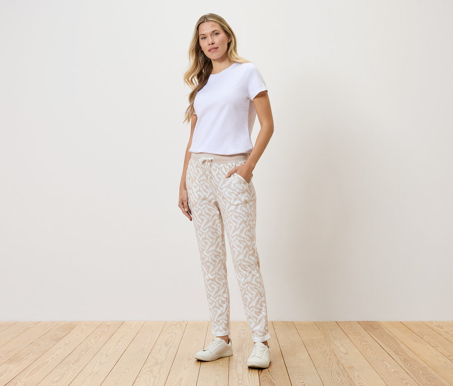 Femme debout devant un mur, portant un t-shirt blanc, un pantalon à motifs et des baskets blanches.