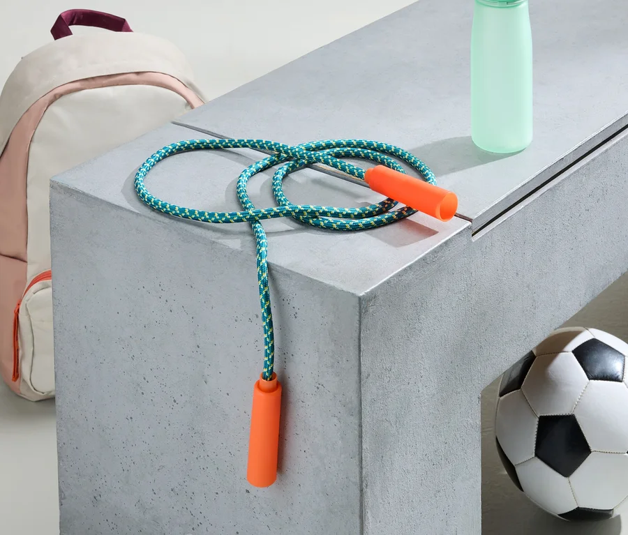 Un sac à dos rose et blanc, une corde à sauter, une bouteille d'eau et un ballon de foot sont posés sur un banc gris.