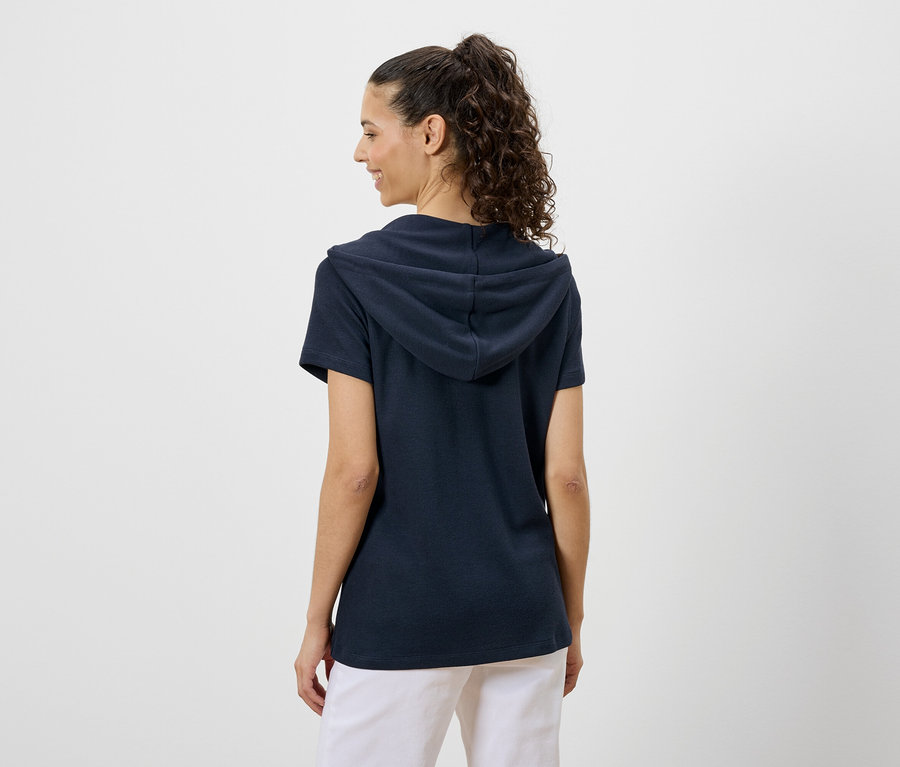 Une femme aux cheveux bouclés porte un t-shirt à capuche bleu marine et un pantalon blanc, vue de dos.