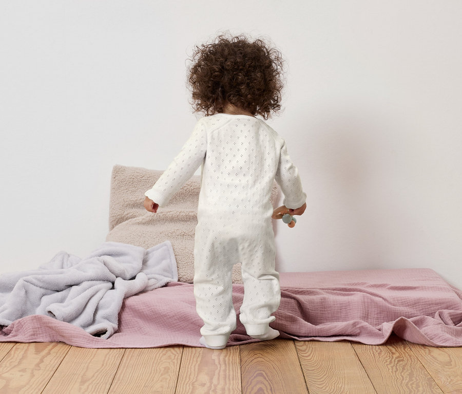 Un jeune enfant aux cheveux bouclés se tient debout sur une couverture rose, à côté d'une couverture grise et d'un coussin beige.