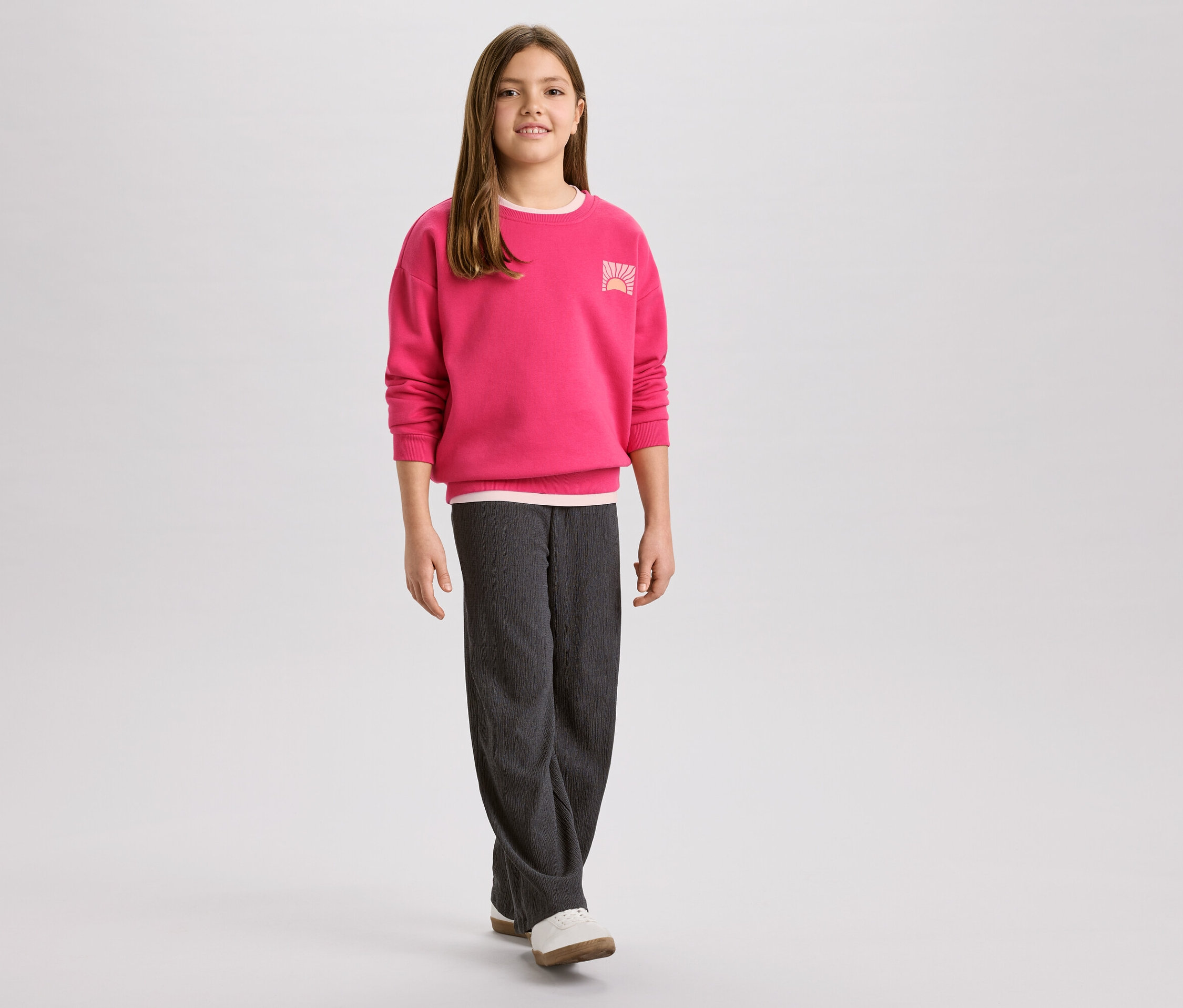Fillette portant un pull rose avec un logo de lever de soleil et un pantalon en velours côtelé gris, avec des chaussures blanches.