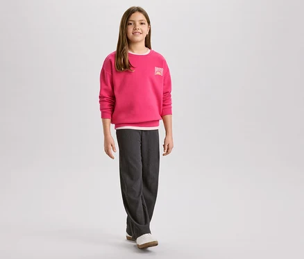 Fillette portant un pull rose avec un logo de lever de soleil et un pantalon en velours côtelé gris, avec des chaussures blanches.