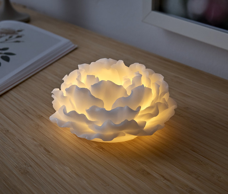 Une lampe blanche en forme de fleur illumine une table en bois. Un livre ouvert est posé à côté.