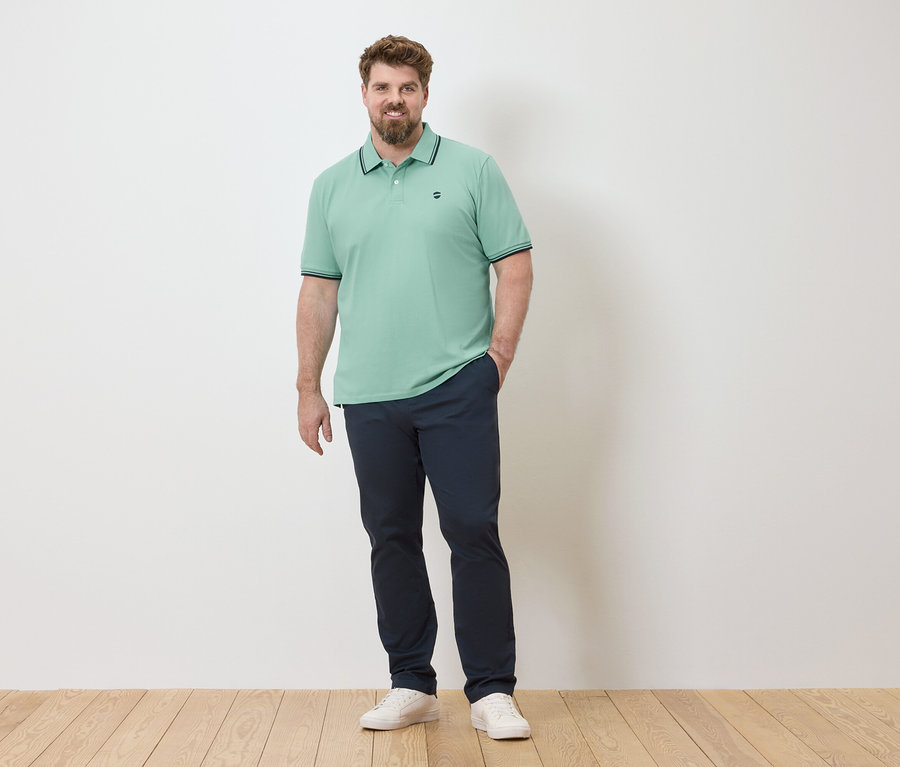 Un homme se tient devant un mur blanc, portant un polo vert clair, un pantalon bleu marine et des baskets blanches.