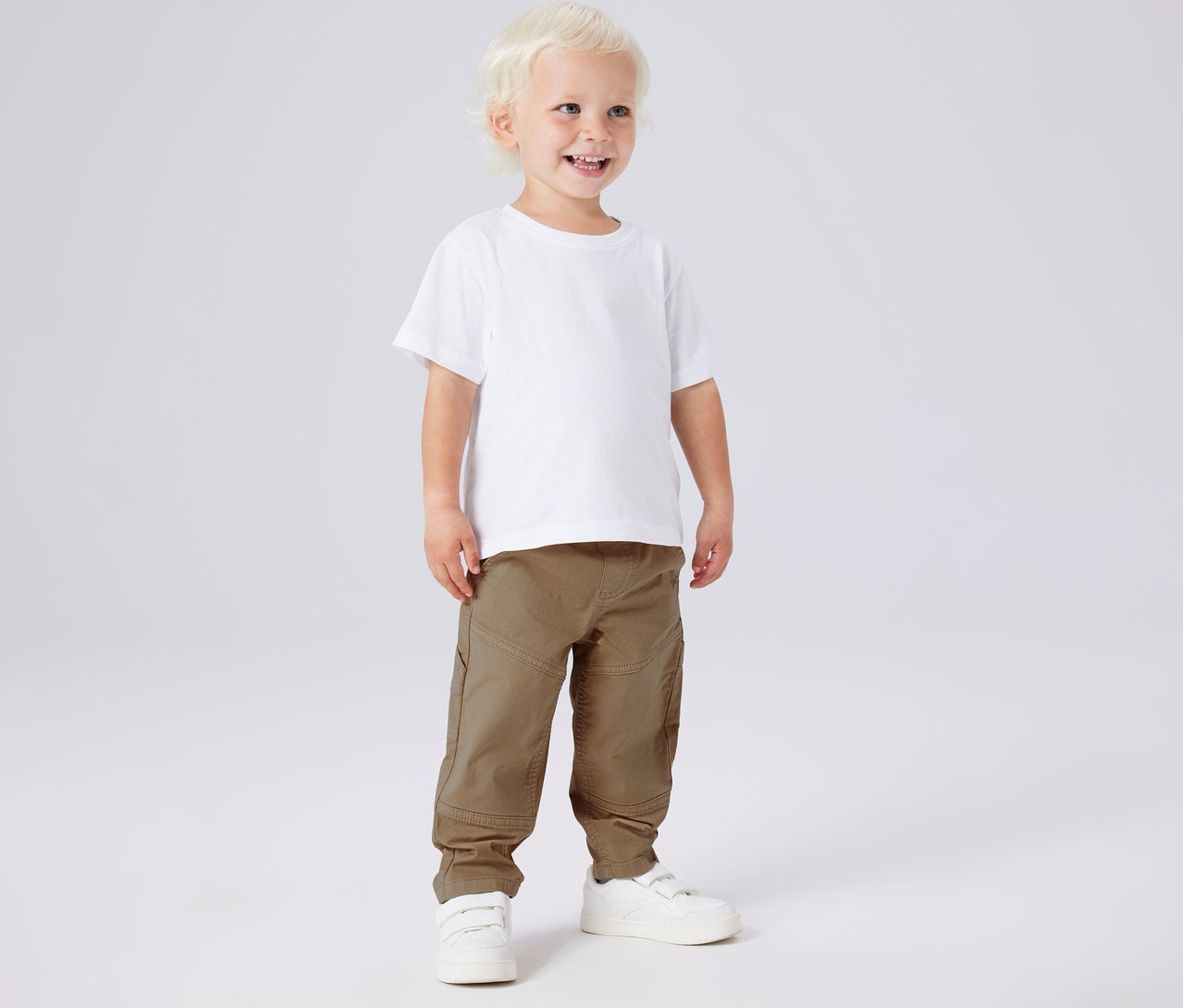 Garçon souriant portant un t-shirt blanc, un pantalon en sergé kaki NAME IT et des chaussures blanches.