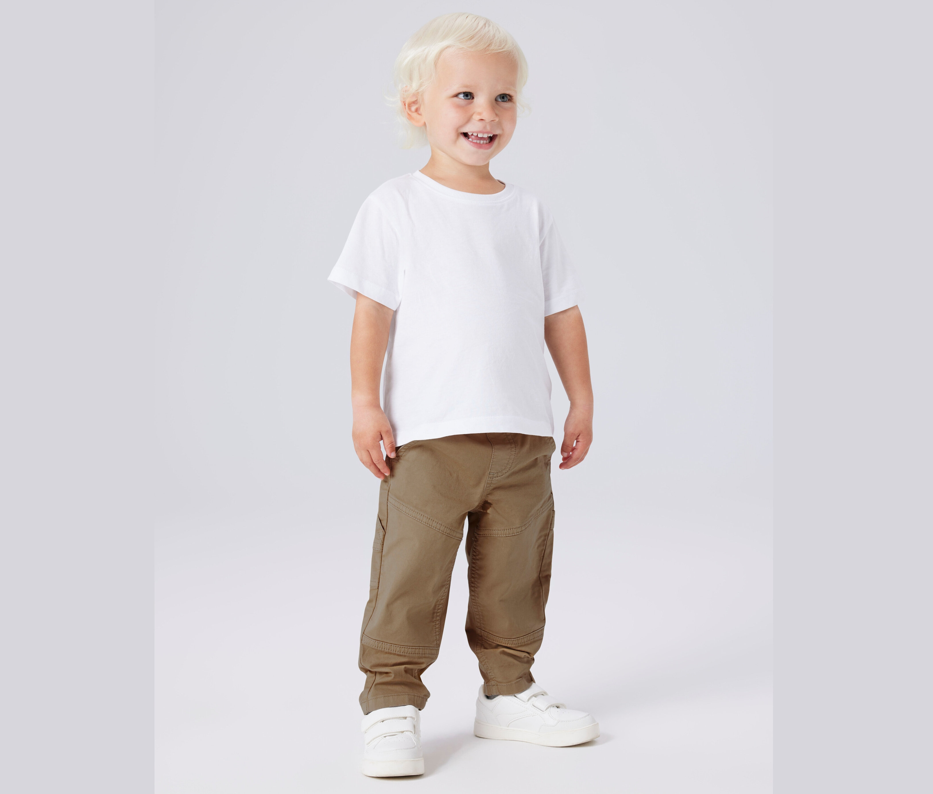 Un jeune garçon blond souriant se tient sur un fond gris vêtu d'un t-shirt blanc, d'un pantalon kaki et de baskets blanches.