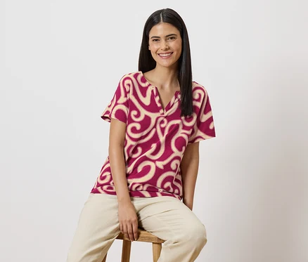 Une femme souriante aux longs cheveux foncés est assise sur un tabouret, portant un chemisier rouge à manches courtes avec un motif beige et un pantalon beige.