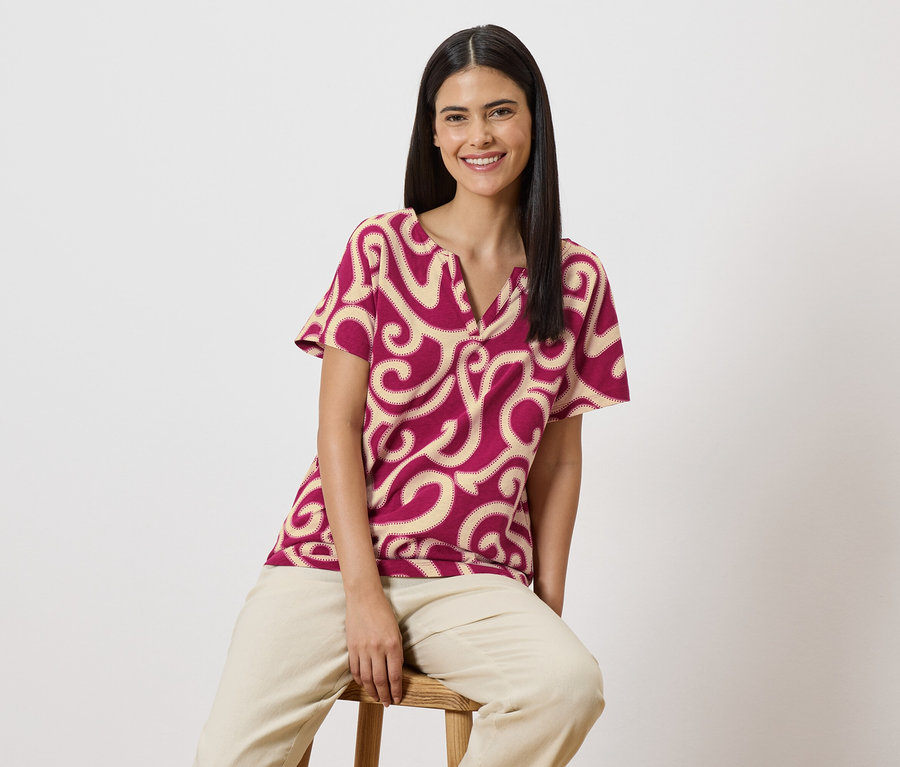 Une femme souriante aux longs cheveux foncés est assise sur un tabouret, portant un chemisier rouge à manches courtes avec un motif beige et un pantalon beige.