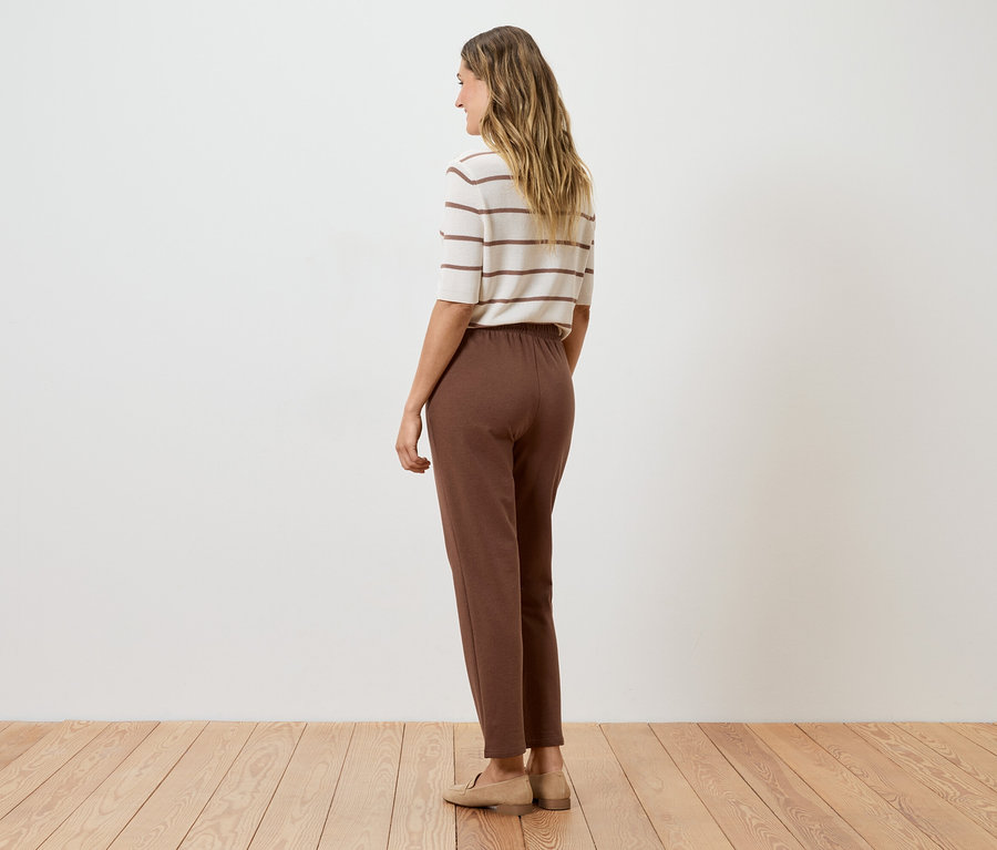Femme vue de dos portant un pantalon marron, un haut rayé et des chaussures beiges devant un mur blanc.