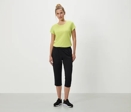 Une femme se tient debout sur un fond blanc, portant un t-shirt vert, un pantacourt noir et des baskets noires.