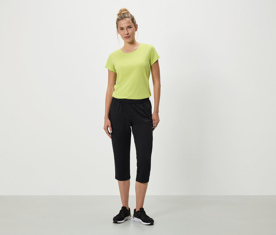 Une femme se tient debout sur un fond blanc, portant un t-shirt vert, un pantacourt noir et des baskets noires.