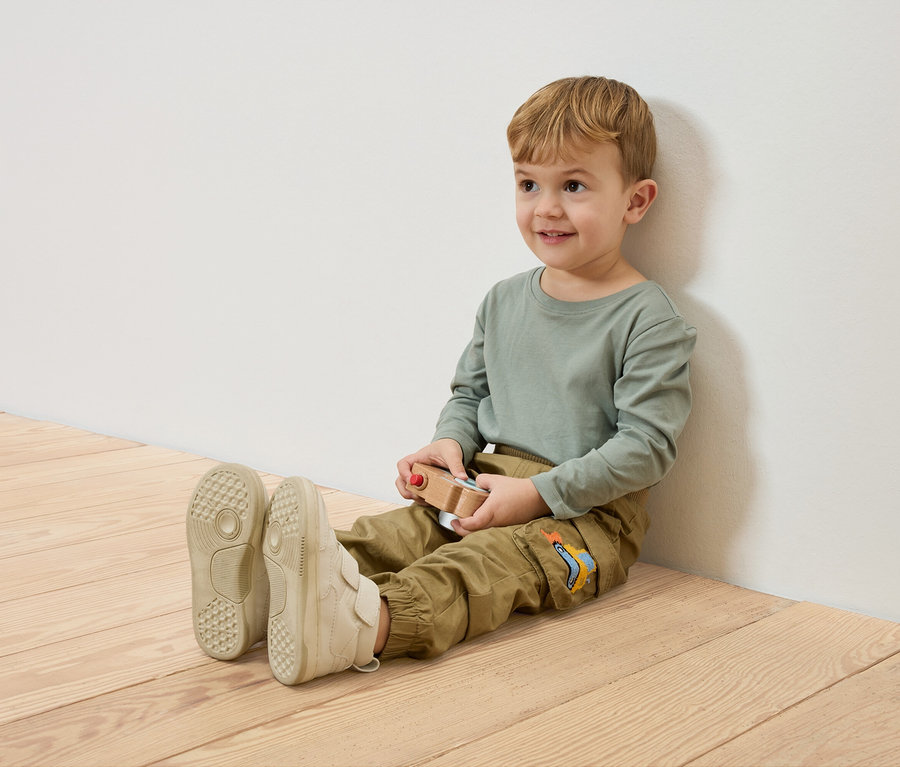 Un garçon souriant est assis sur le plancher en bois, appuyé contre un mur blanc, tenant une voiture jouet en bois.