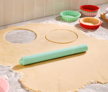 Sur un plan de travail fariné, une pâte étalée avec deux cercles découpés, un rouleau à pâtisserie vert et des moules à tartelettes colorés.