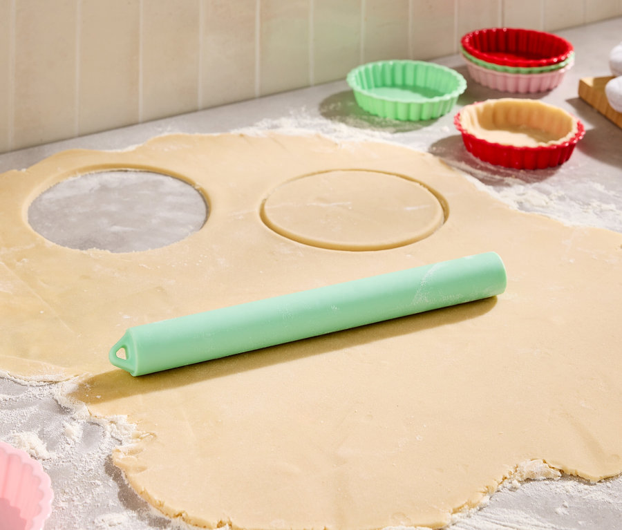 Sur un plan de travail fariné, une pâte étalée avec deux cercles découpés, un rouleau à pâtisserie vert et des moules à tartelettes colorés.