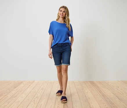 Une femme se tient devant un mur blanc, portant un t-shirt bleu, un short en jean et des sandales.