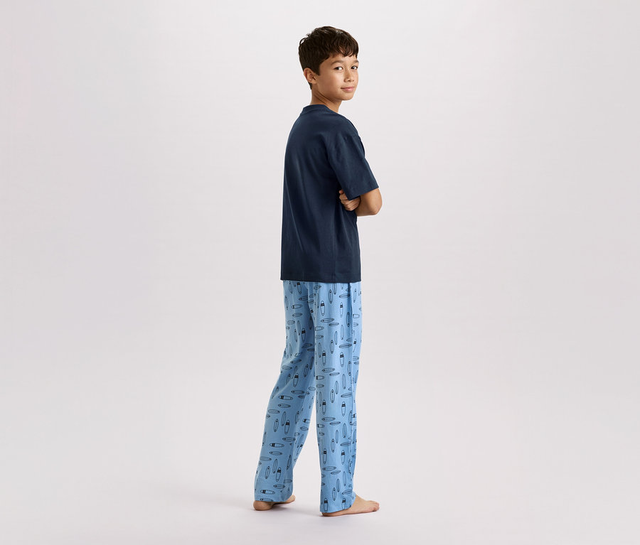 Un garçon se tient les bras croisés, portant un t-shirt bleu marine et un pantalon de pyjama bleu clair avec un motif de planches de surf.