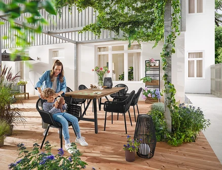 Une mère et son enfant sont assis à une table de jardin extensible avec un cadre rustique d'environ 200 à 250 cm. Une petite et une grande Lanterne solaire avec ruban en tissu sont posées sur le sol. À l'arrière-plan, Mur végétal avec étagères flexiblement accrochables « Elin ».