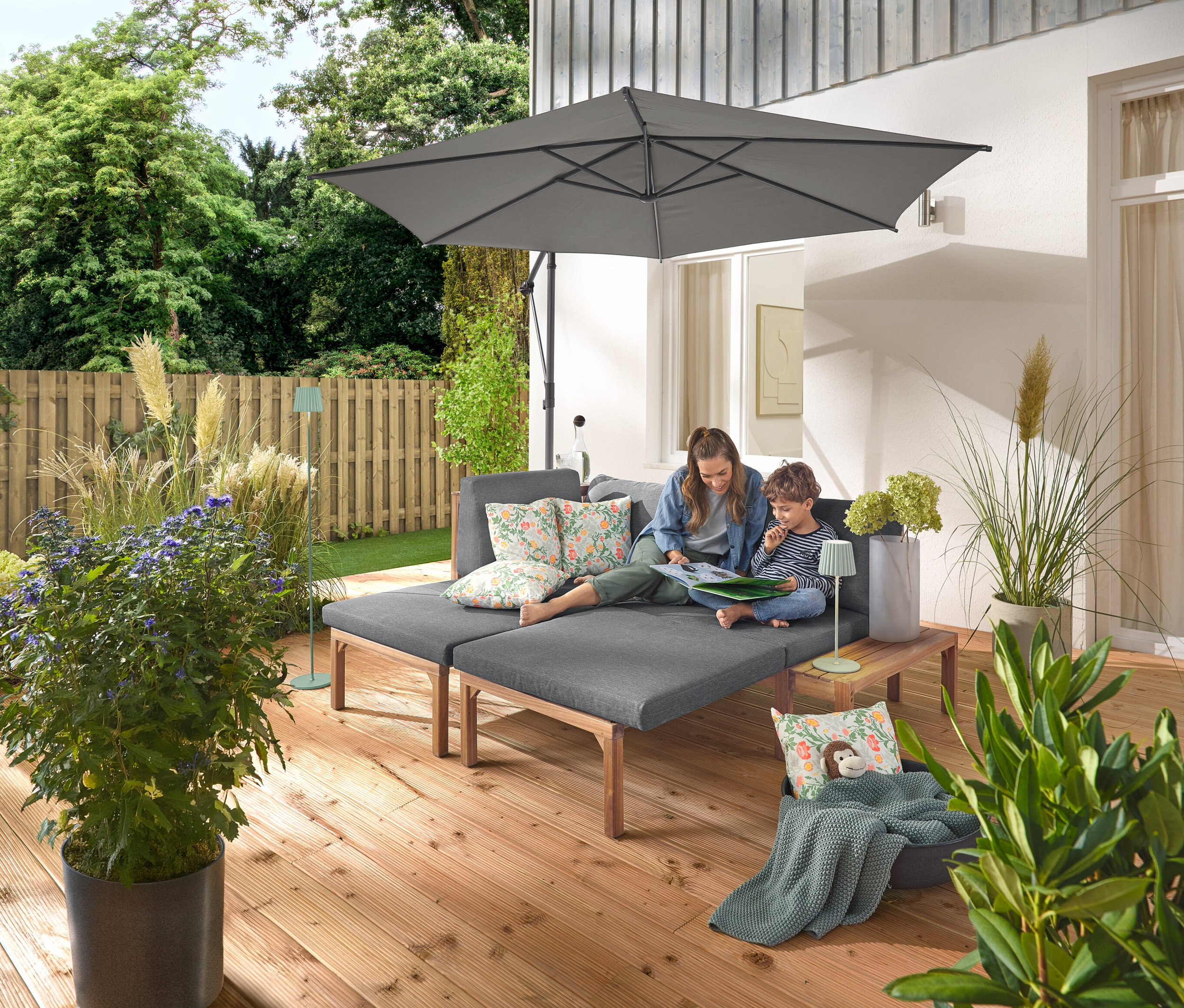 Une femme et un enfant sont assis sur un canapé de jardin gris avec des repose-pieds extensibles séparément. Deux coussins d’extérieur avec motif floral sont posés sur le canapé. Un parasol Leco anthracite avec pied et housse offre de l’ombre. Une lampe de table d’extérieur à LED rechargeable sauge est posée sur une table en bois à côté du canapé, tandis qu’un lampadaire d’extérieur à LED rechargeable sauge est visible à l’arrière-plan.
