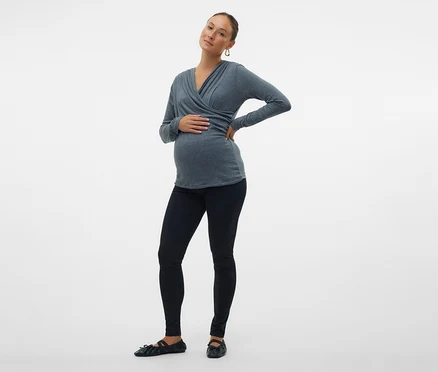 Femme enceinte posant sur un fond blanc, portant un haut cache-cœur gris, un legging noir et des ballerines.