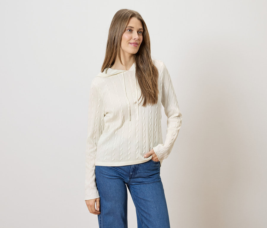 Une femme aux longs cheveux bruns porte un sweat à capuche torsadé crème et un jean bleu.