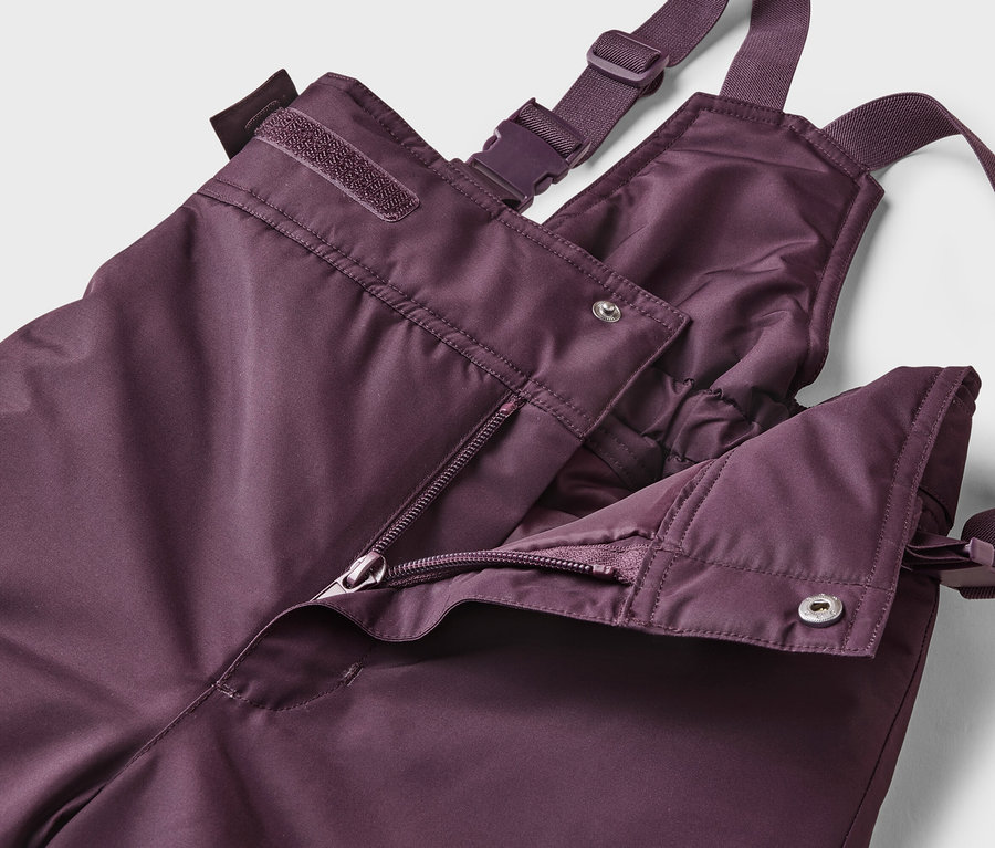 Gros plan d'un pantalon de ski aubergine ouvert à bretelles avec fermeture éclair et boutons sur fond blanc.