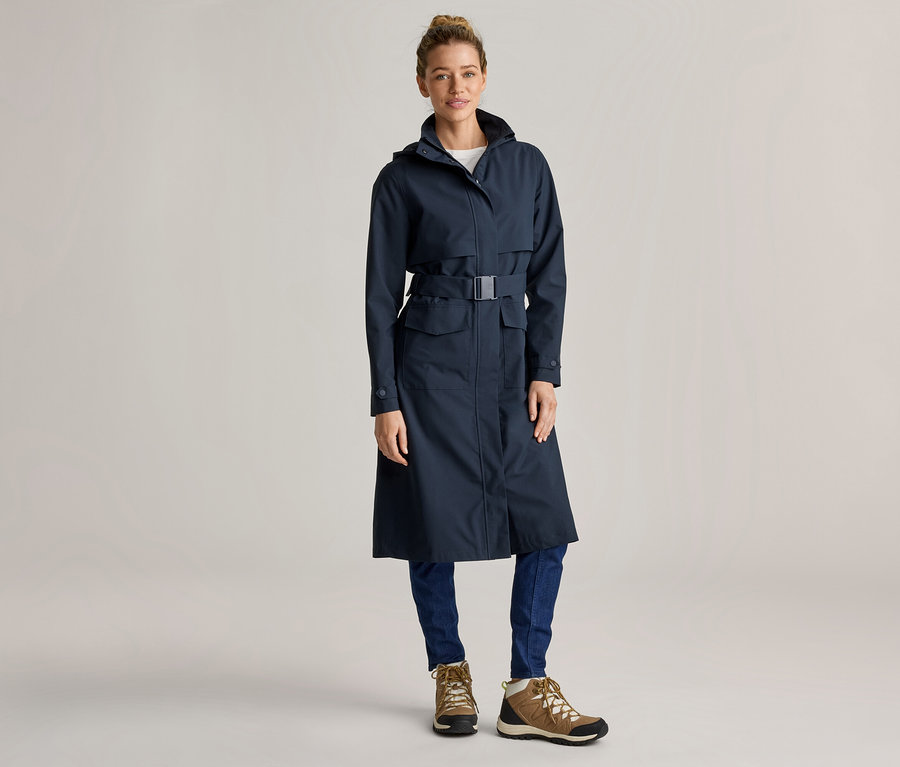 Une femme portant un chignon porte un long imperméable bleu marine ceinturé, un jean bleu et des chaussures de randonnée marron sur un fond gris clair.