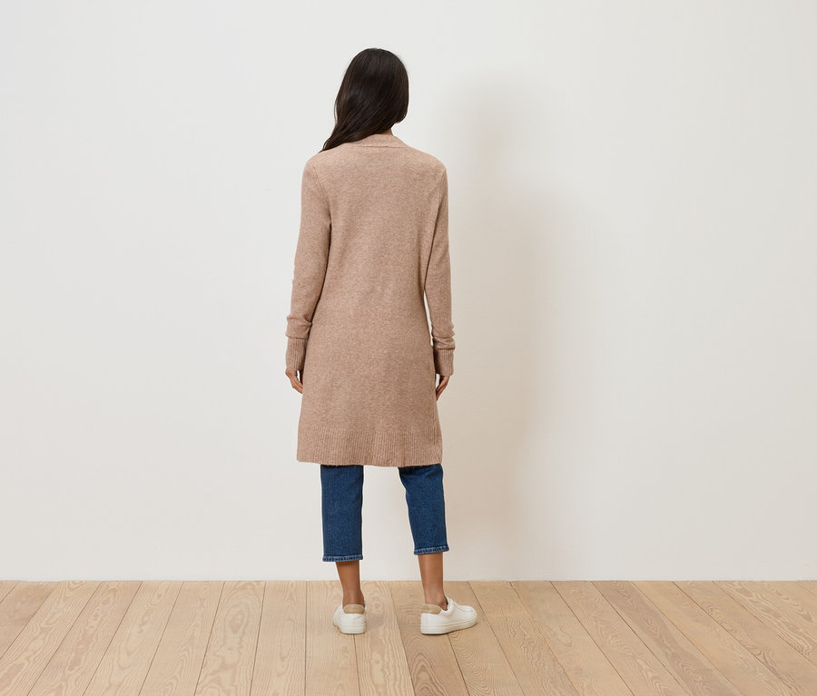 Vue de dos d'une femme aux longs cheveux foncés portant un long cardigan beige, un jean bleu et des chaussures blanches.