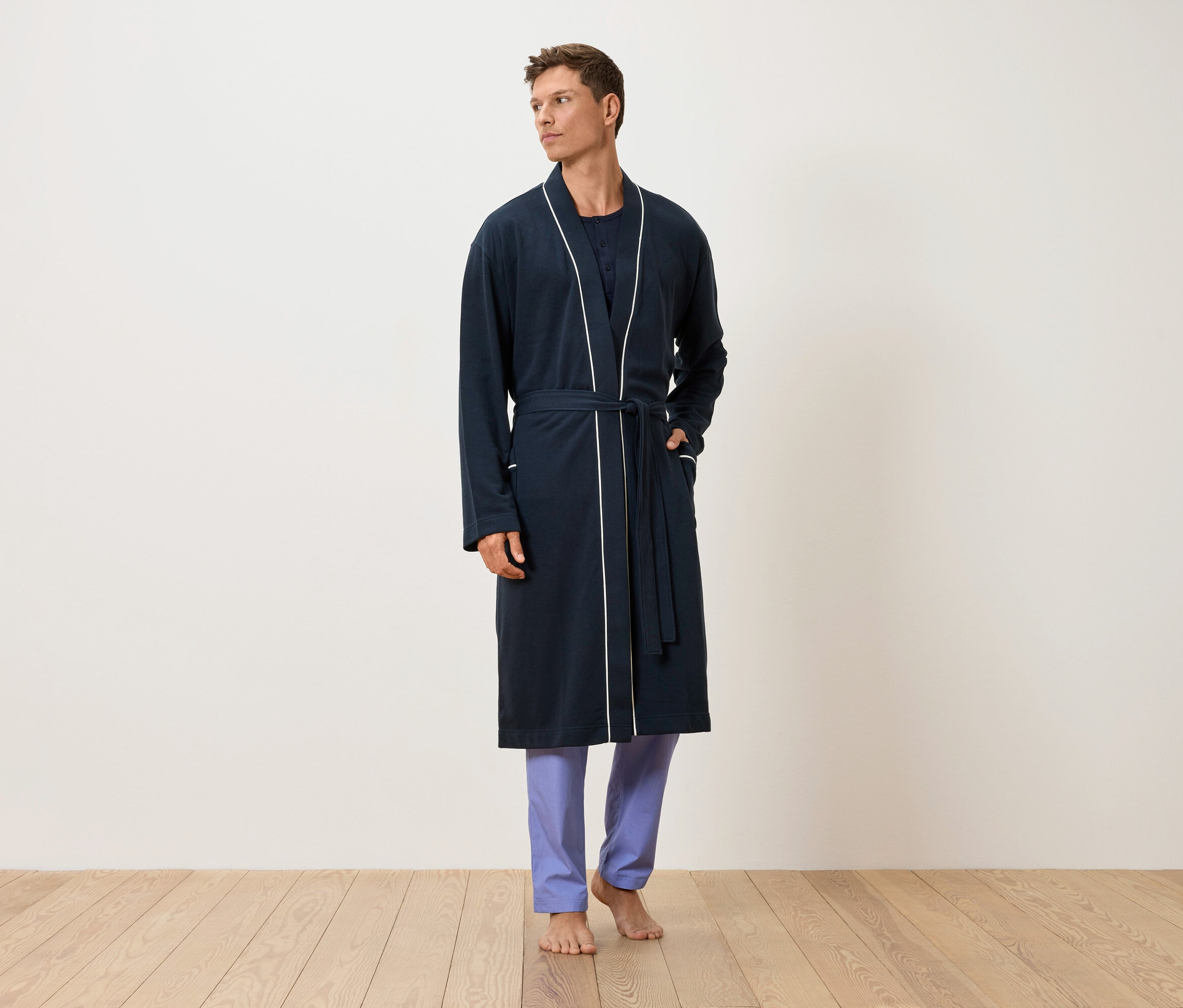 Un homme se tient pieds nus sur un plancher en bois, vêtu d'un peignoir bleu marine avec des finitions blanches et d'un pantalon bleu.
