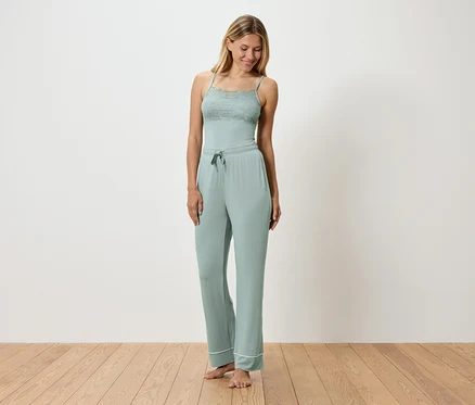 Une femme porte un pyjama vert clair avec des détails en dentelle, debout sur un plancher en bois devant un mur blanc.