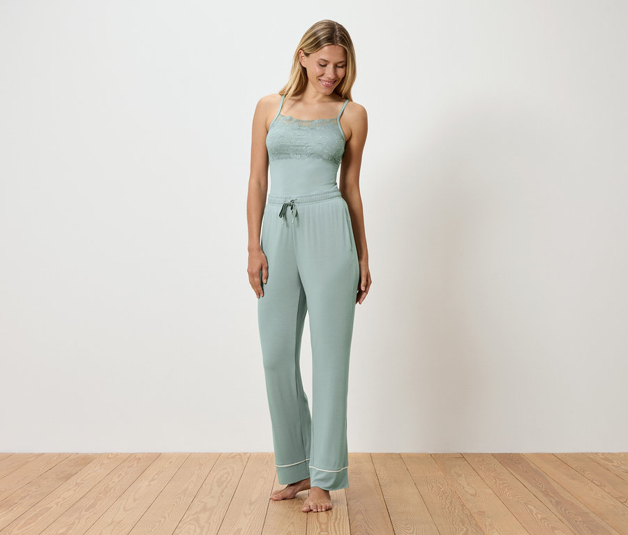 Une femme porte un pyjama vert clair avec des détails en dentelle, debout sur un plancher en bois devant un mur blanc.