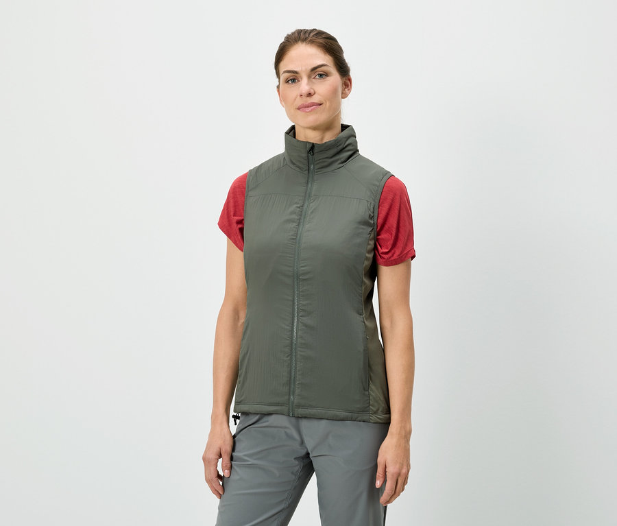 Une femme debout porte un gilet vert olive sur un t-shirt rouge, associé à un pantalon gris.