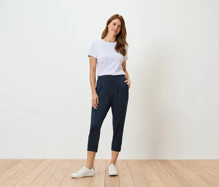 Femme en t-shirt blanc et pantalon bleu marine debout devant un mur blanc.