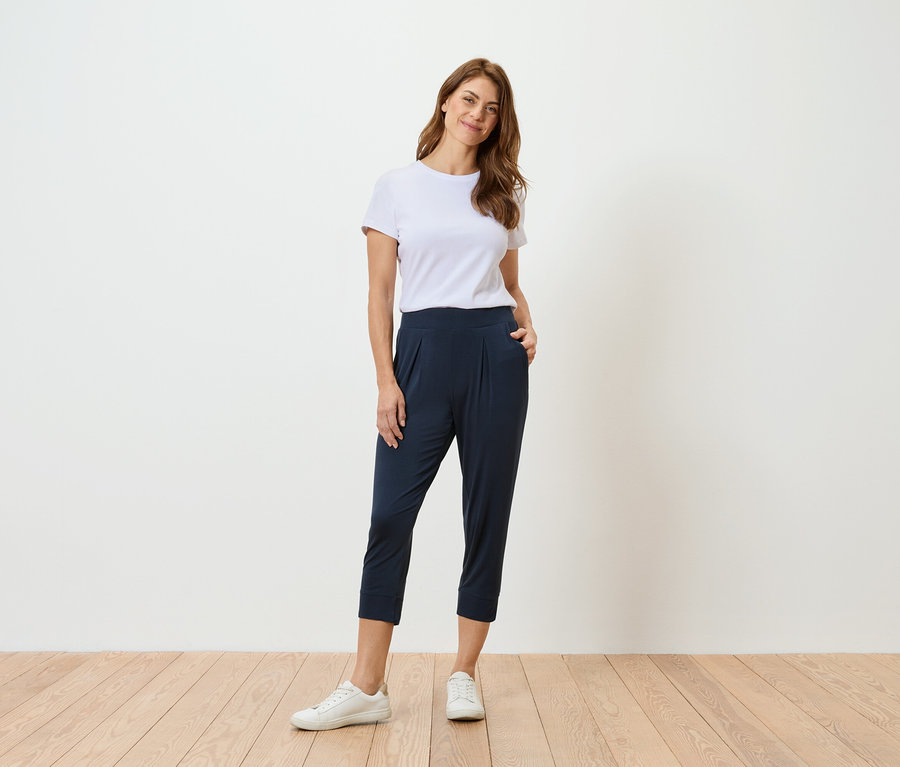 Femme en t-shirt blanc et pantalon bleu marine debout devant un mur blanc.