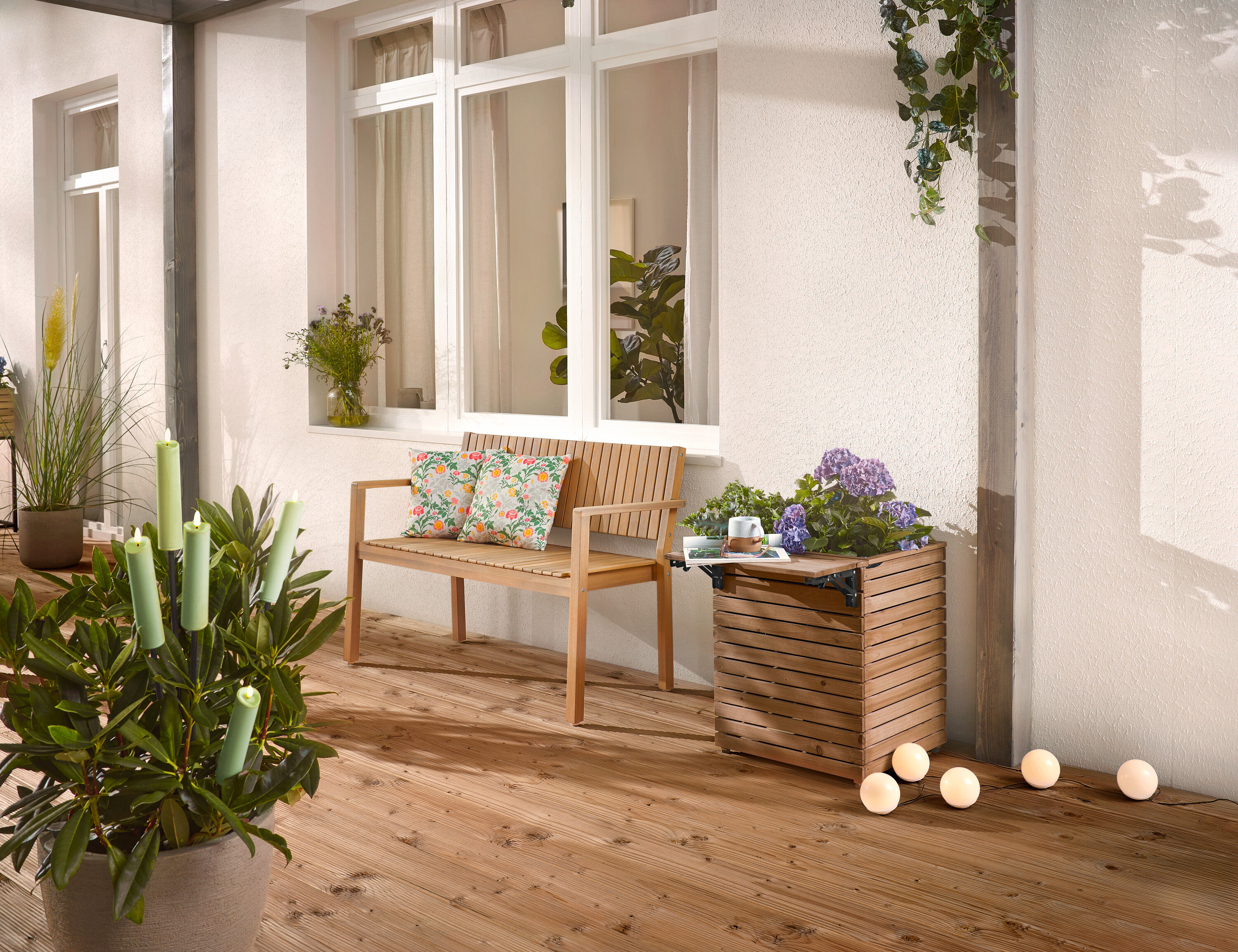 Sur une terrasse en bois se trouvent un banc en bois avec des coussins, un bac à plantes avec des fleurs et une plante en pot avec des bougies.