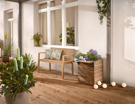Sur une terrasse en bois se trouvent un banc en bois avec des coussins, un bac à plantes avec des fleurs et une plante en pot avec des bougies.