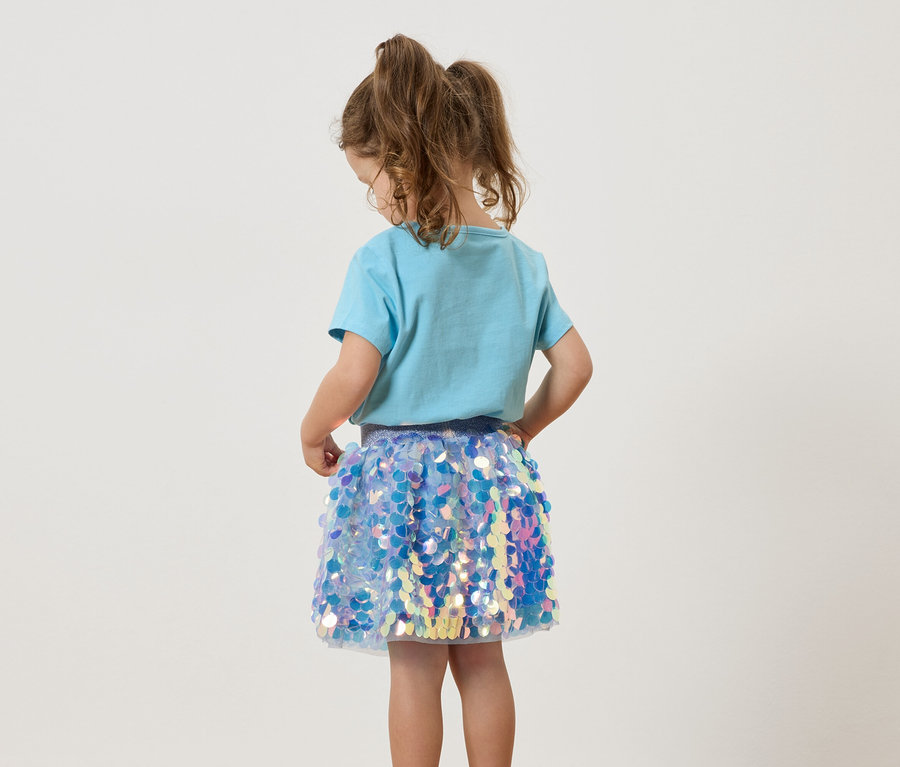 Une jeune fille avec des tresses porte un t-shirt bleu clair et une jupe à paillettes scintillantes.