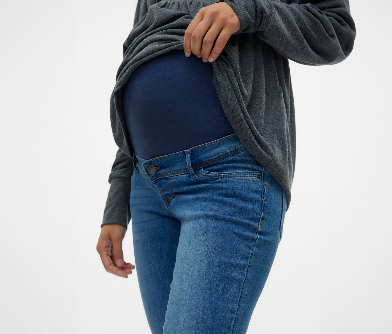 Femme remontant un haut gris au-dessus de son ventre, portant un Jeans Skinny MAMALICIOUS «
MLMILA » et un haut de maternité bleu marine.
