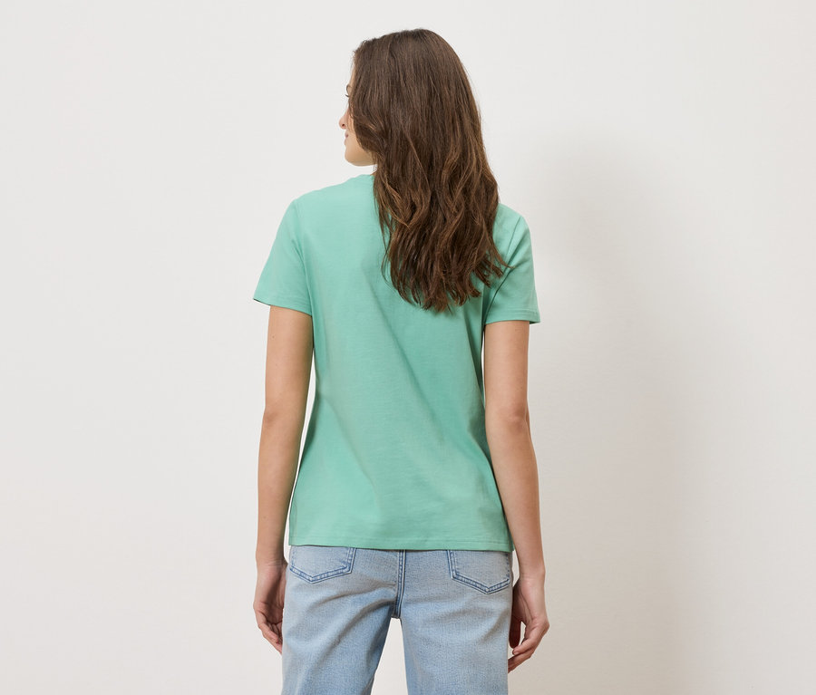 Une femme aux longs cheveux bruns porte un t-shirt vert clair et un jean bleu.