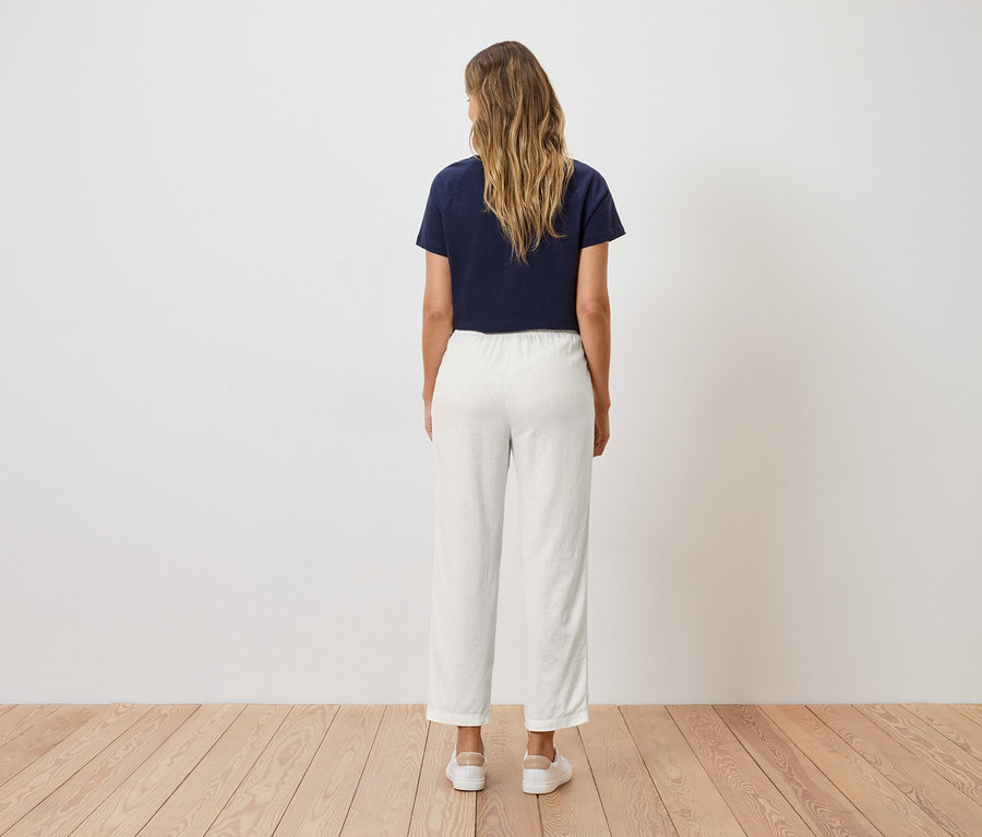 Une femme aux cheveux blonds et ondulés se tient dos au spectateur, portant un t-shirt bleu marine et un pantalon blanc.