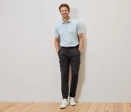 Un homme en polo bleu clair et pantalon cargo gris se tient devant un mur, les mains dans les poches.