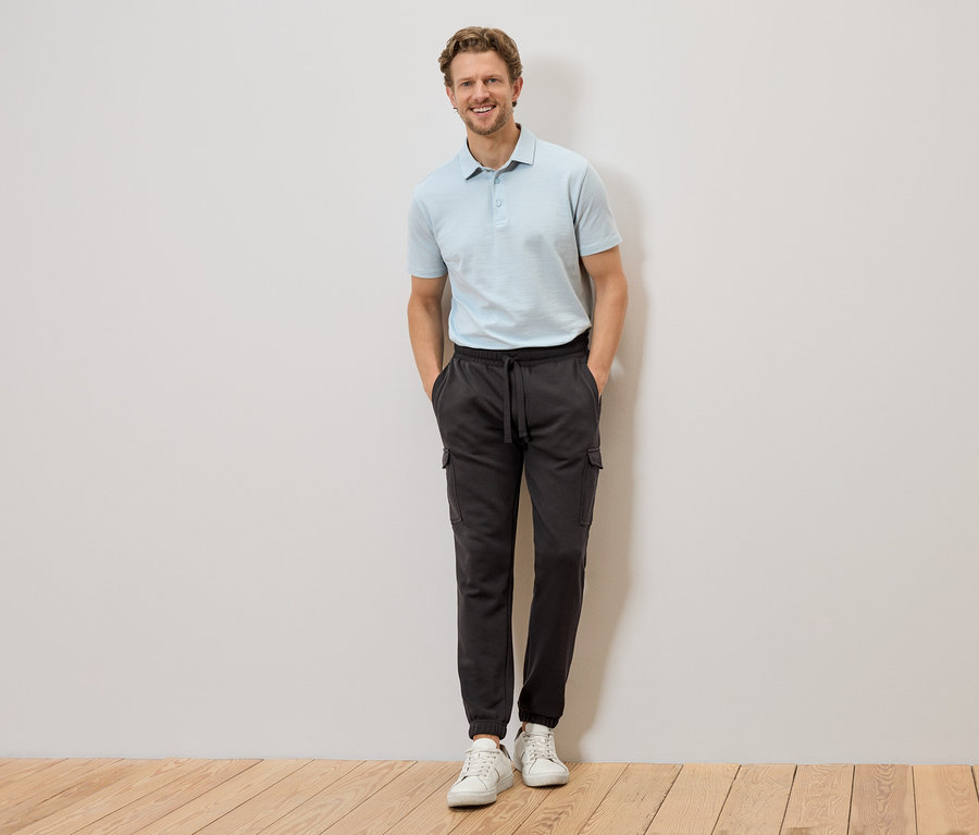Un homme en polo bleu clair et pantalon cargo gris se tient devant un mur, les mains dans les poches.