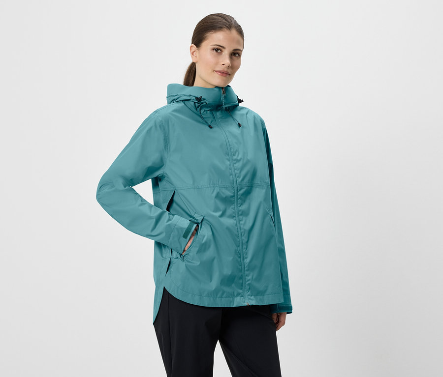 Femme portant une veste à capuche bleu pétrole, la main dans la poche, et un pantalon noir sur fond blanc.