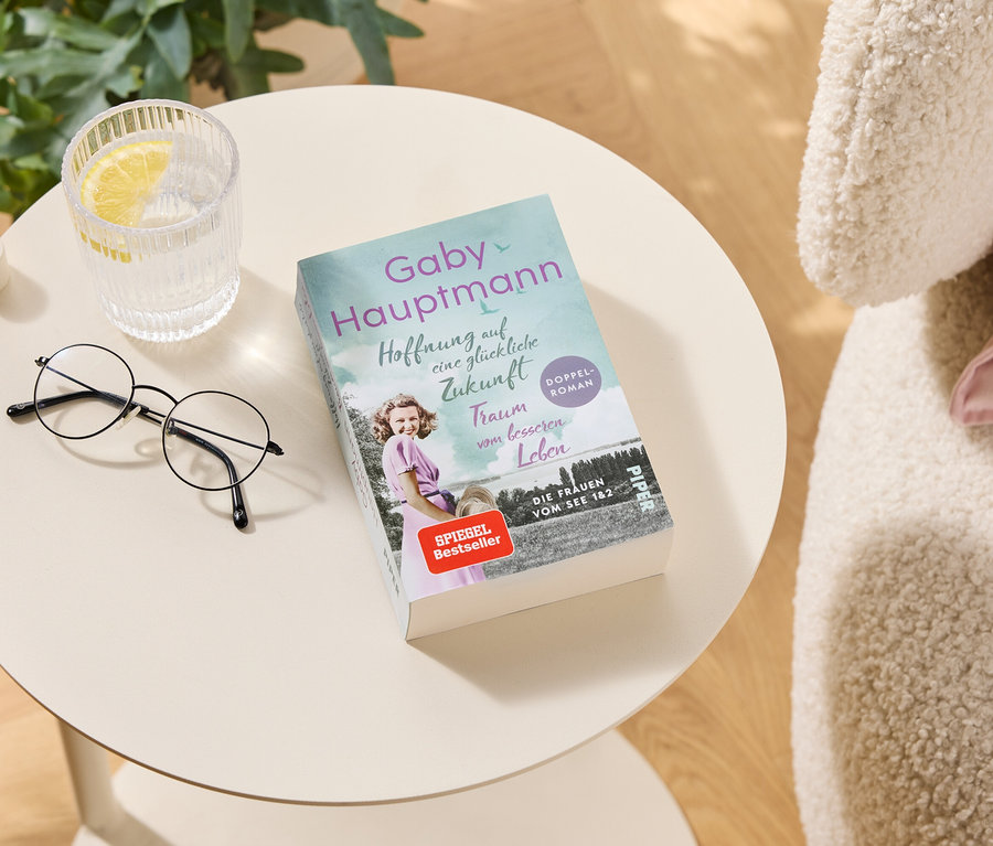 Sur une table, il y a des lunettes, un verre d'eau avec du citron et le livre "Hoffnung auf eine glückliche Zukunft" de Gaby Hauptmann.