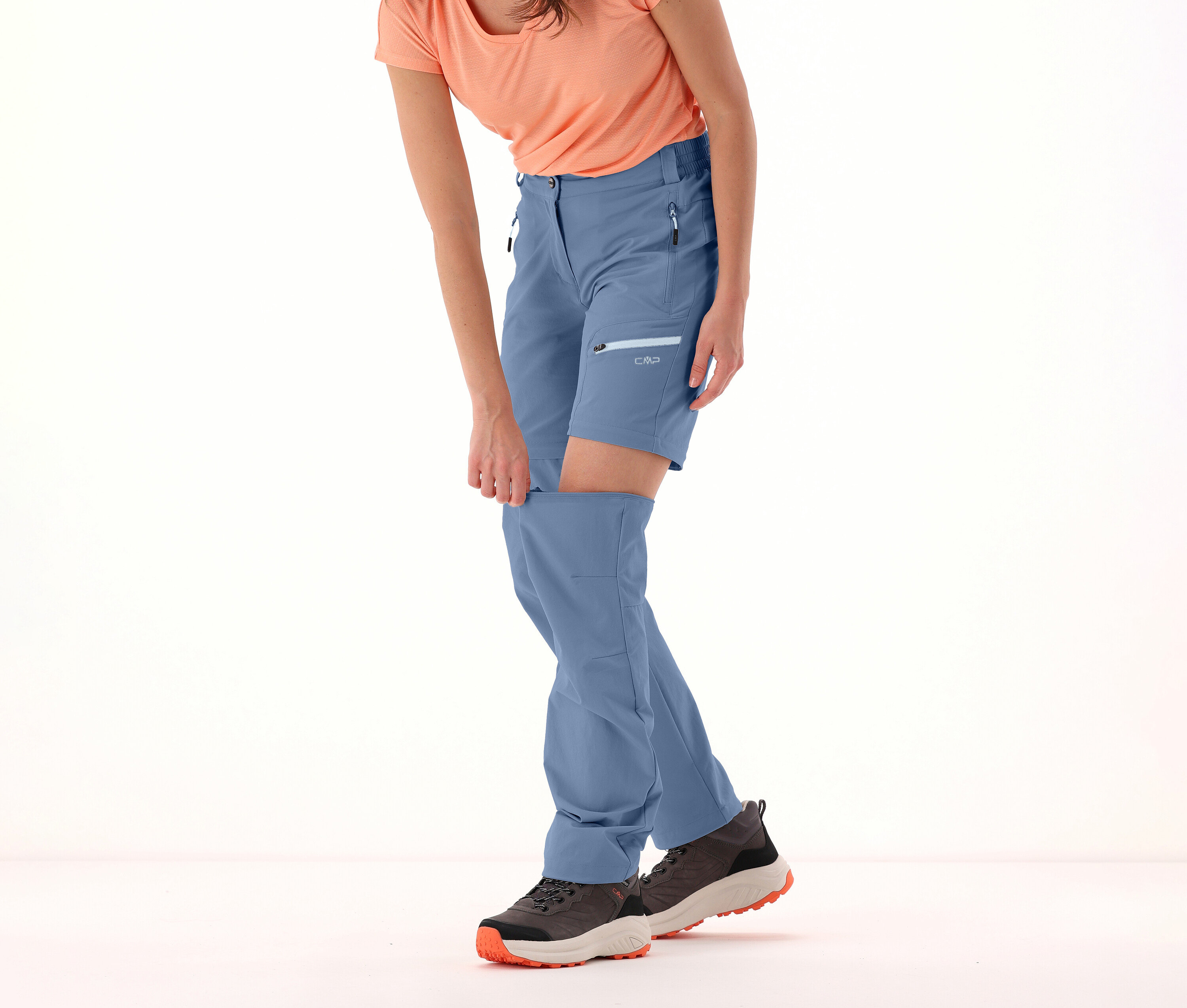 Femme dézippant les jambes du pantalon zip-off CMP pour femme bleu. Elle porte un t-shirt et des chaussures de randonnée.