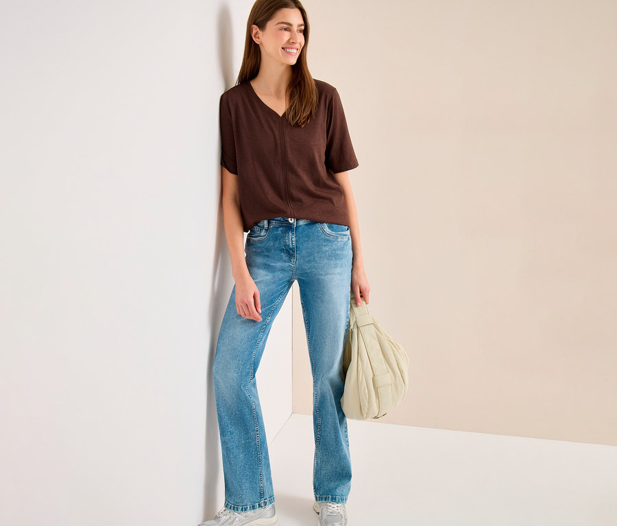 Une femme souriante portant une tunique CECIL marron, un jean évasé bleu clair et des baskets argentées, est appuyée contre un mur blanc et tient un sac à main matelassé beige clair.