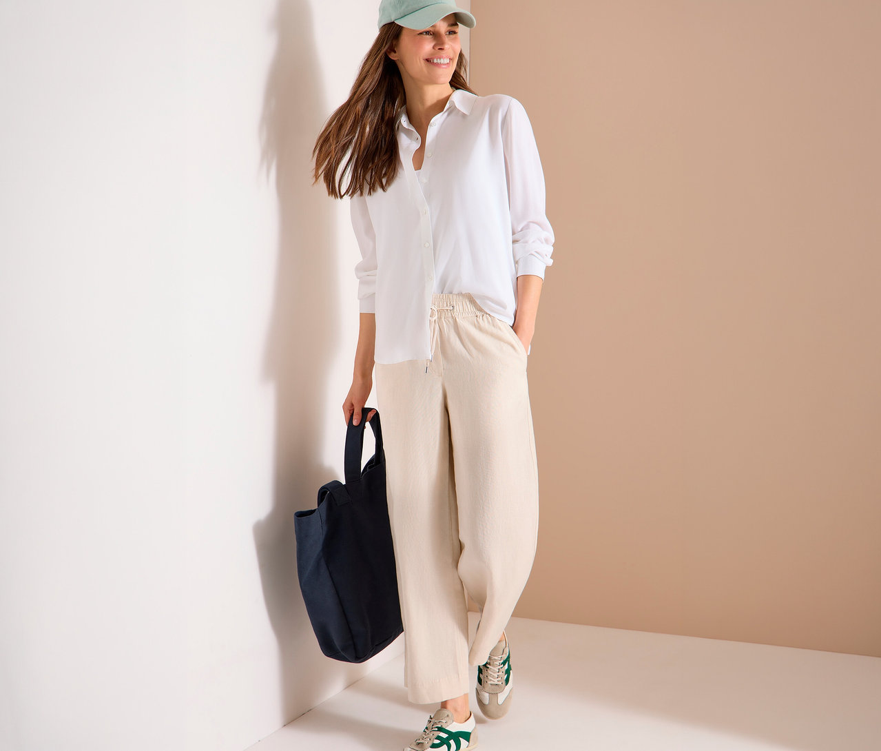 Une femme portant une casquette vert clair, une chemise blanche, un pantalon beige et des baskets tient un sac en tissu bleu marine.