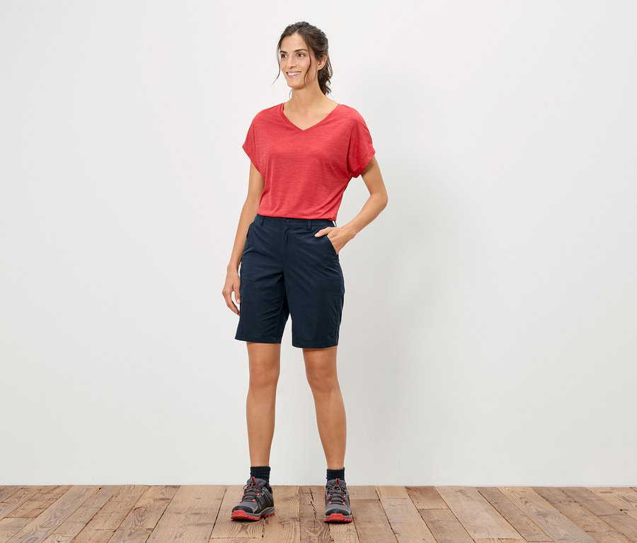 Une femme se tient debout, la main dans la poche, portant un t-shirt rouge et un short bleu.